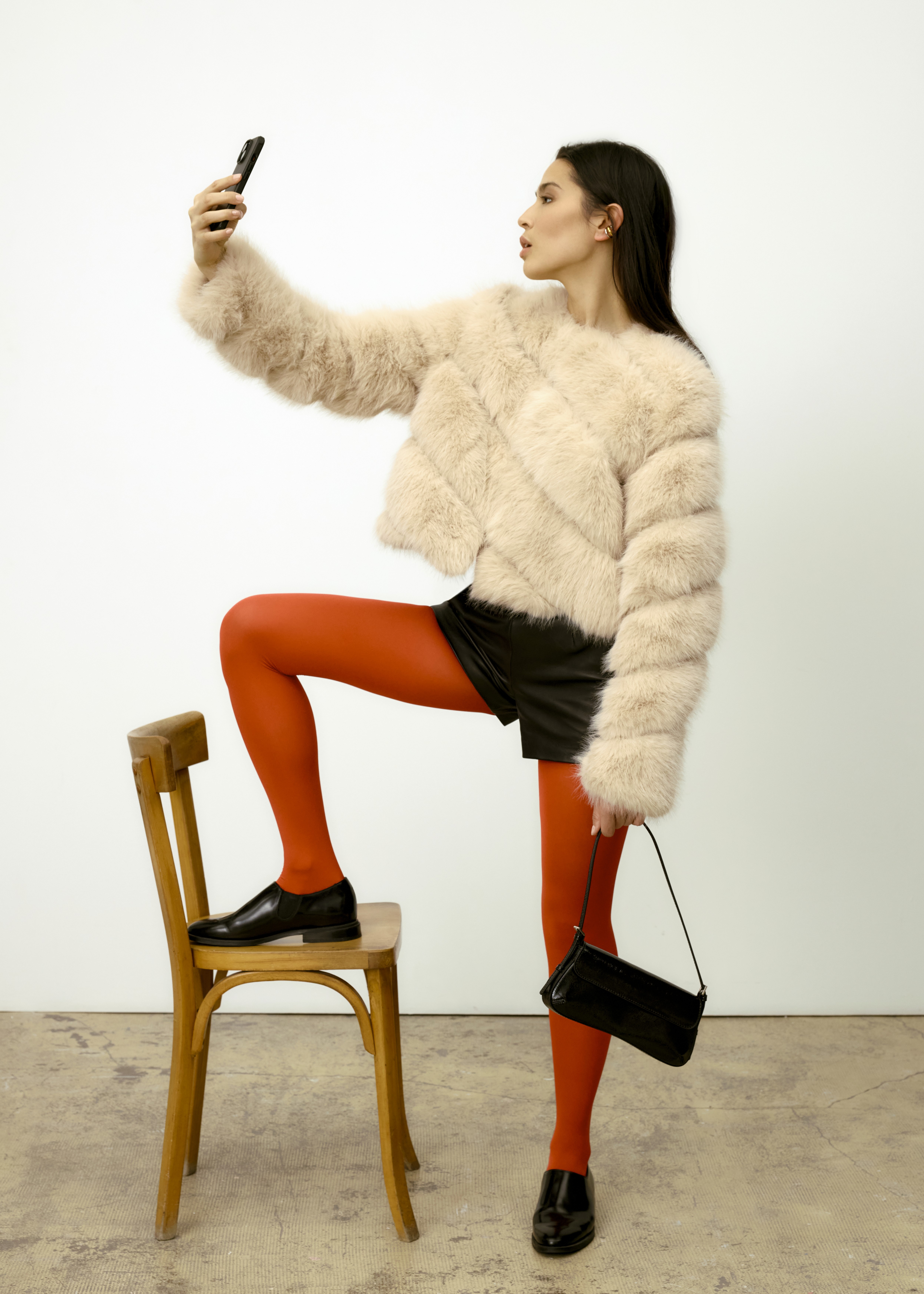 Femme prenant un selfie, un pied posé sur une chaise, portant une veste claire et des collants rouges