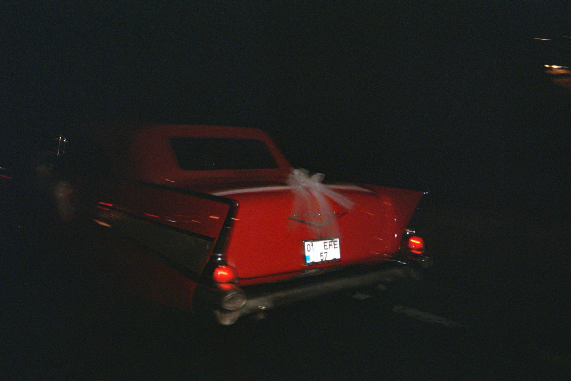 A vintage red car with a white ribbon tied to the back is driving at night, conveying a sense of nostalgia and celebration on a dimly lit street.