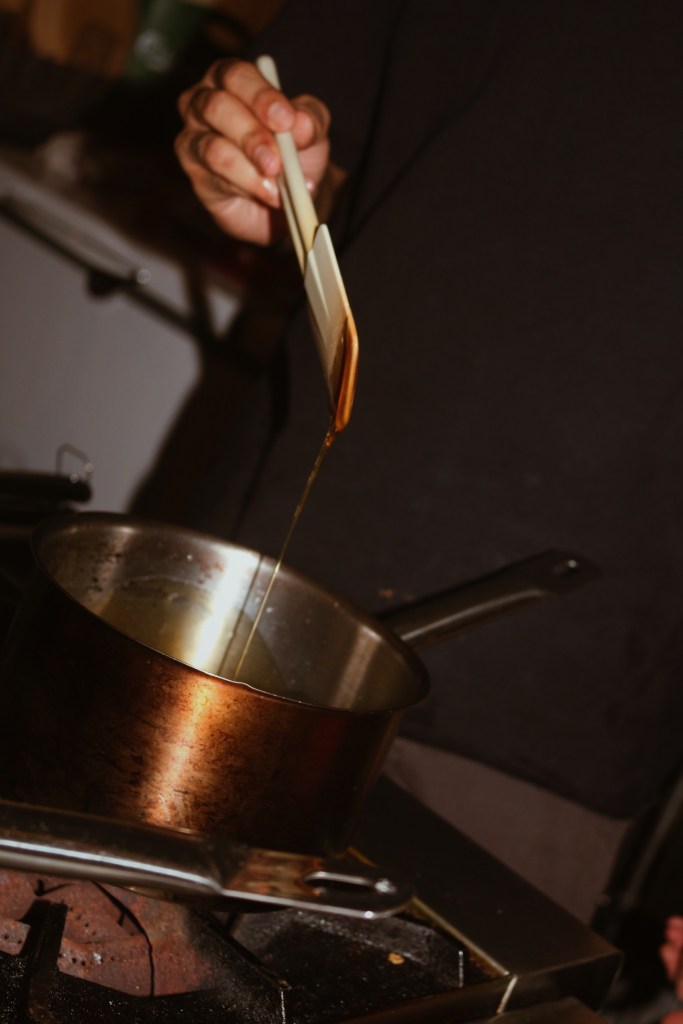 A person's hand holding a spatula over a pot on a stovetop, with liquid being poured from the spatula into the pot.