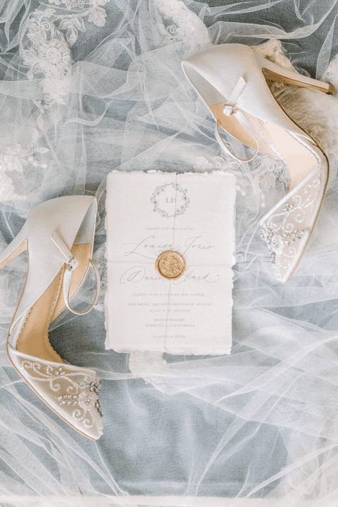 Artistic flat-lay photograph of bridal shoes and an embossed wedding invitation on delicate lace, exemplifying sophisticated, fine art wedding photography with a focus on elegance and detail.