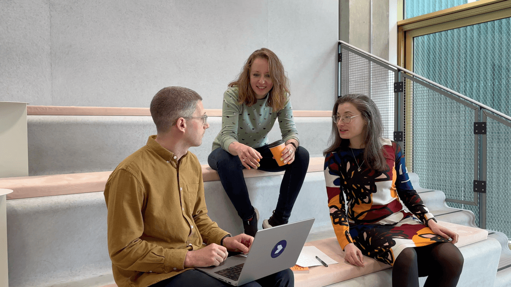 Three designers talking to each other in a meeting room. One has a laptop and one is drinking a coffee.