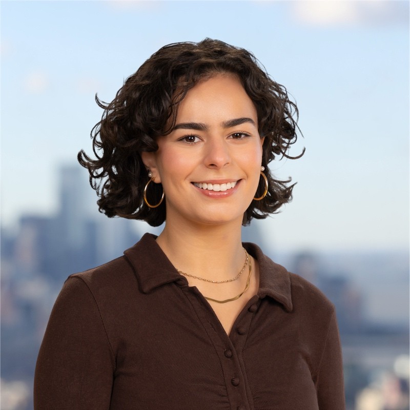 A young woman with long brown hair smiles warmly against a green background, dressed in a black jacket and white shirt.