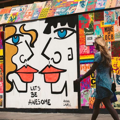 Colorful street art mural featuring abstract faces with bold lines and vibrant colors. A person in a blue dress walks by.