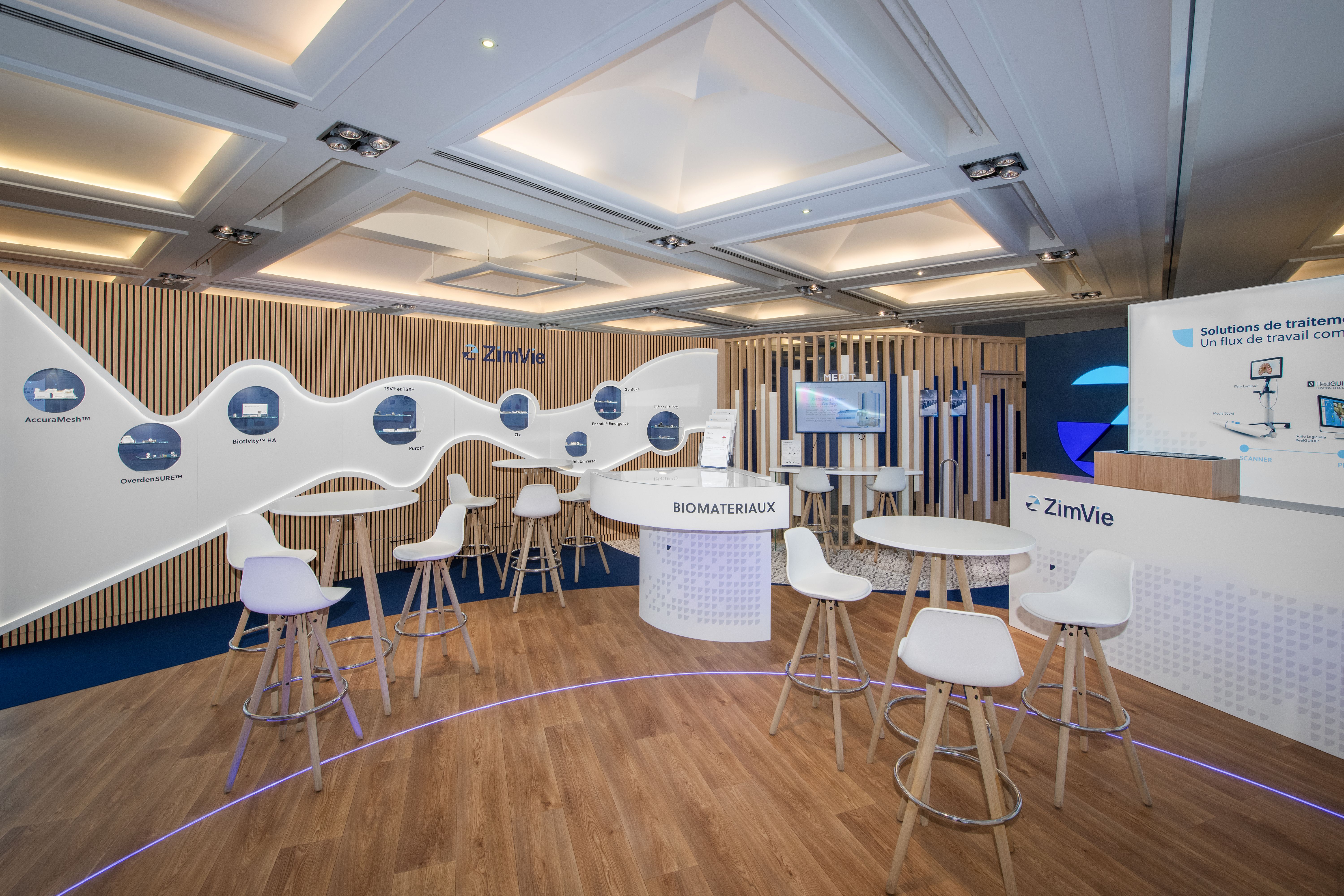Stand ZimVie congrès ADF 2025 réalisé par Expace – vue intérieure avec comptoir Biomatériaux rétroéclairé, frise produits organique en vague, lames bois et espace lounge tabourets blancs parquet