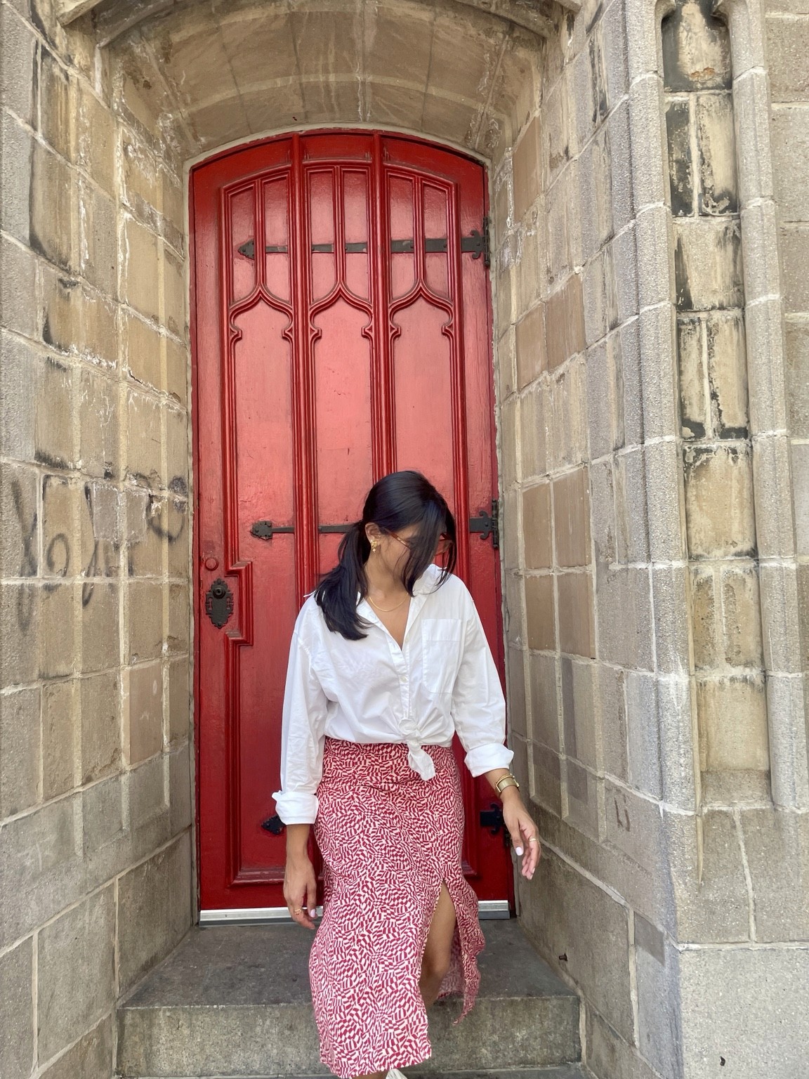 Pranjali in a white blouse and patterned red skirt inside a stone niche with a red door.
