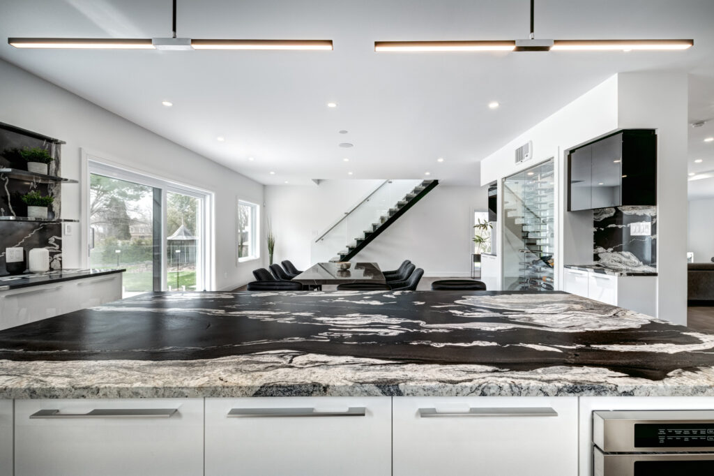Bright open-concept kitchen and dining area with black and white marble waterfall island, sleek white cabinetry, and glass staircase, showcasing B&Y Reno Corp’s custom kitchen remodeling in a modern luxury home.
