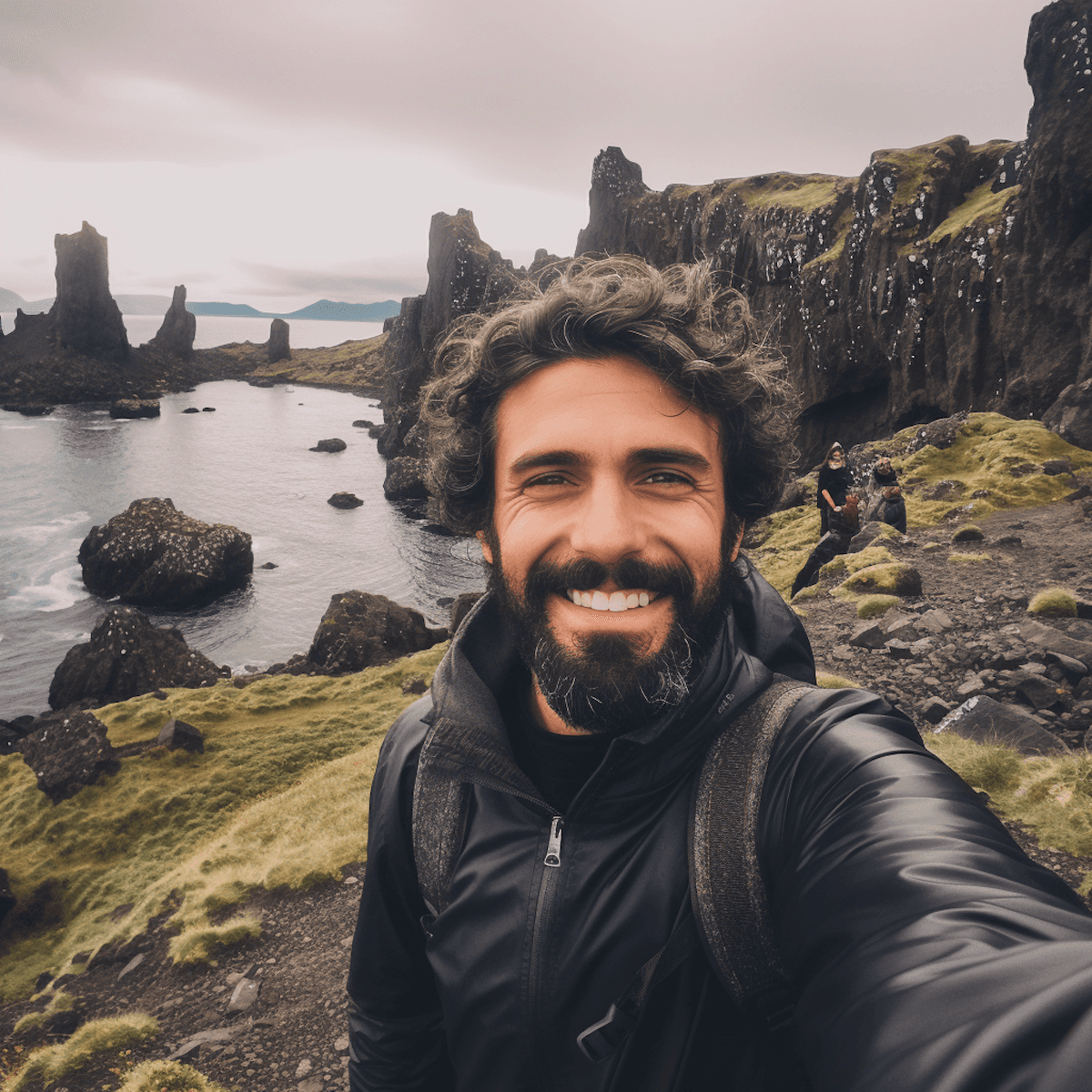 A smiling man with curly hair stands on a rocky coast, with rugged rocks and a cloudy sky in the background.
