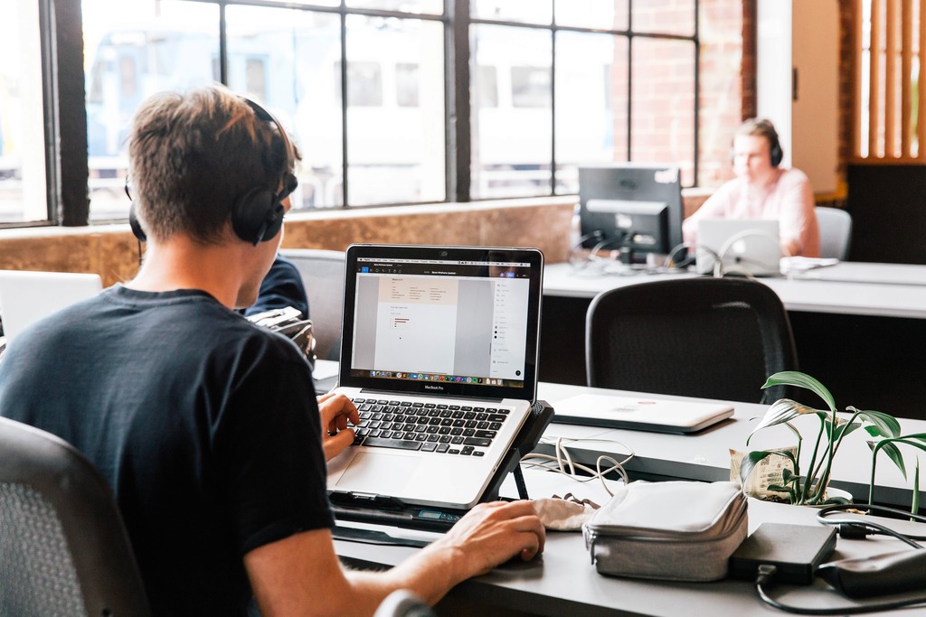 A person wearing headphones works at a laptop in a modern office setting, surrounded by other individuals at their desks, under bright natural light from large windows.