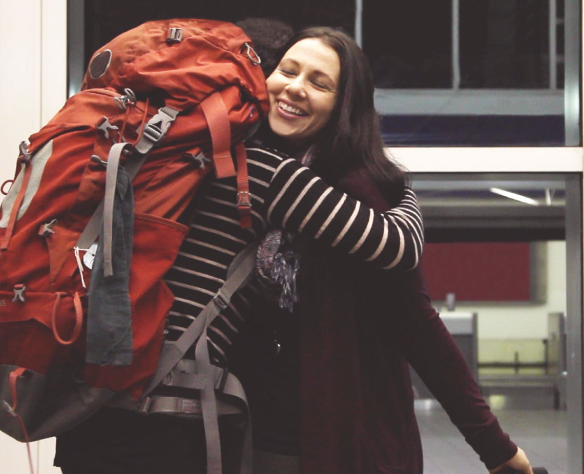 A woman with long dark hair wears a black and white striped long-sleeved shirt and a dark red cardigan. She is hugging a large orange backpack with gray straps and buckles. The backpack is positioned on her back. She is smiling and looking directly at the camera. The background is blurred, showing a glass door or window and a dark interior space.