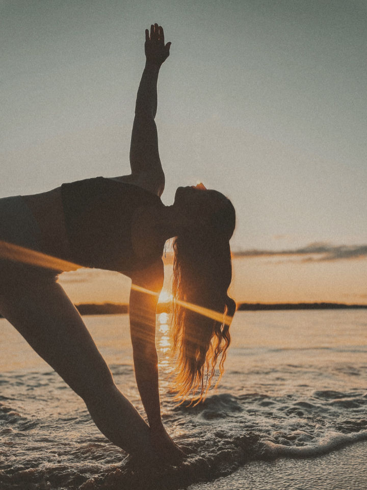 Woman silhouetted in a yoga triangle pose against a golden sunset over calm water