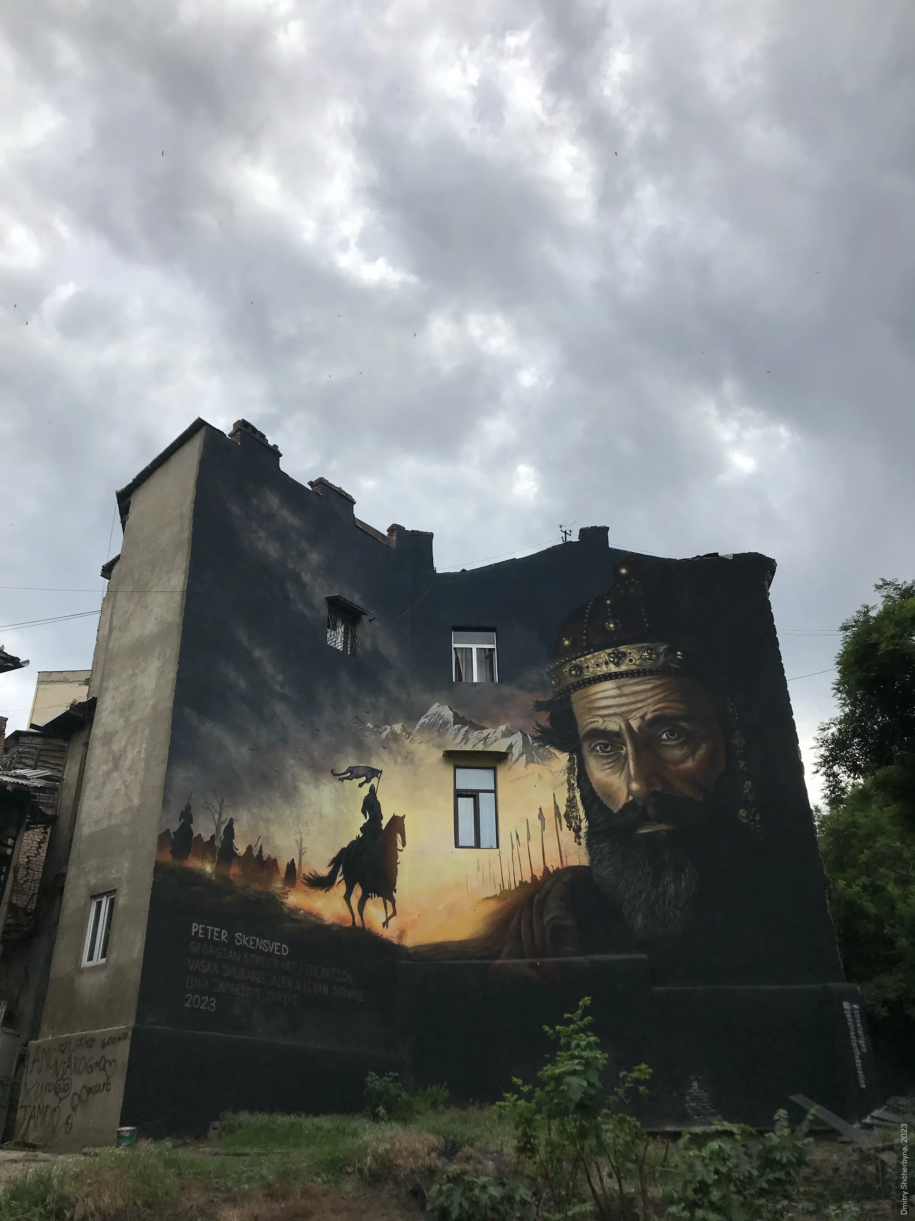 A mural covering the side of an old building shows the close-up face of a bearded king wearing an ornate crown. Next to him is a scene of a horseman carrying a banner in front of snow-capped mountains, with an army of soldiers bearing spears at sunset.