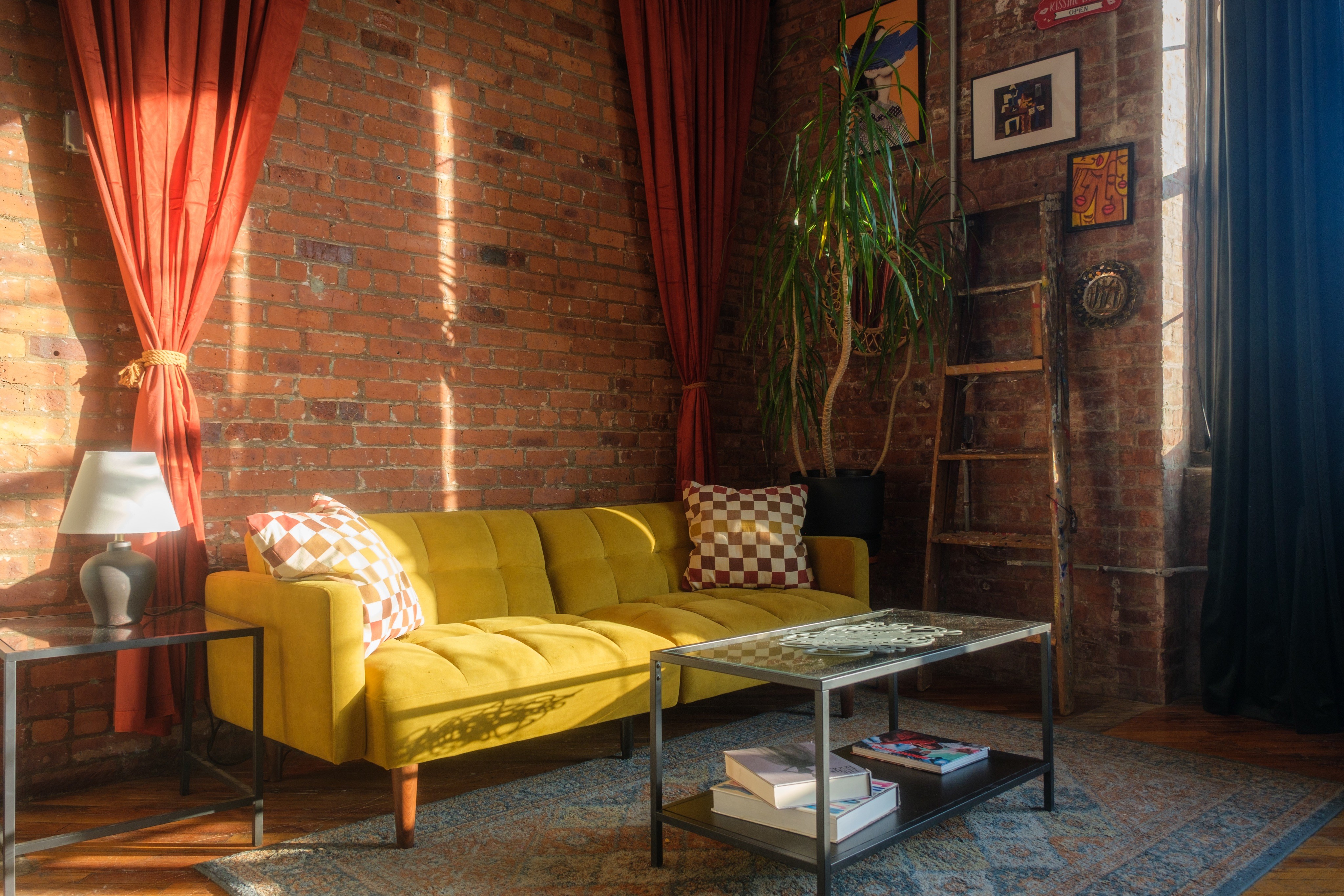 A sunlit room with exposed brick walls features a yellow couch with checkered pillows, vibrant plants, and eclectic decor, creating a cozy and inviting atmosphere. Natural light streams in from a large window with yellow curtains, adding warmth to the space.