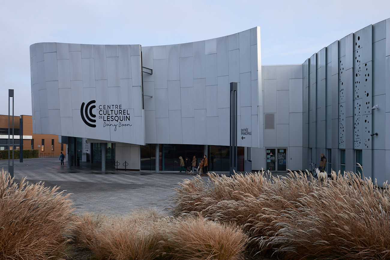 Vue de face, entrée du centre culturel de Lesquin avec l'inscription du lettrage noir sur béton blanc, quelque humains au abord de l'entré et quelques hautes herbes orangées au premier plan.