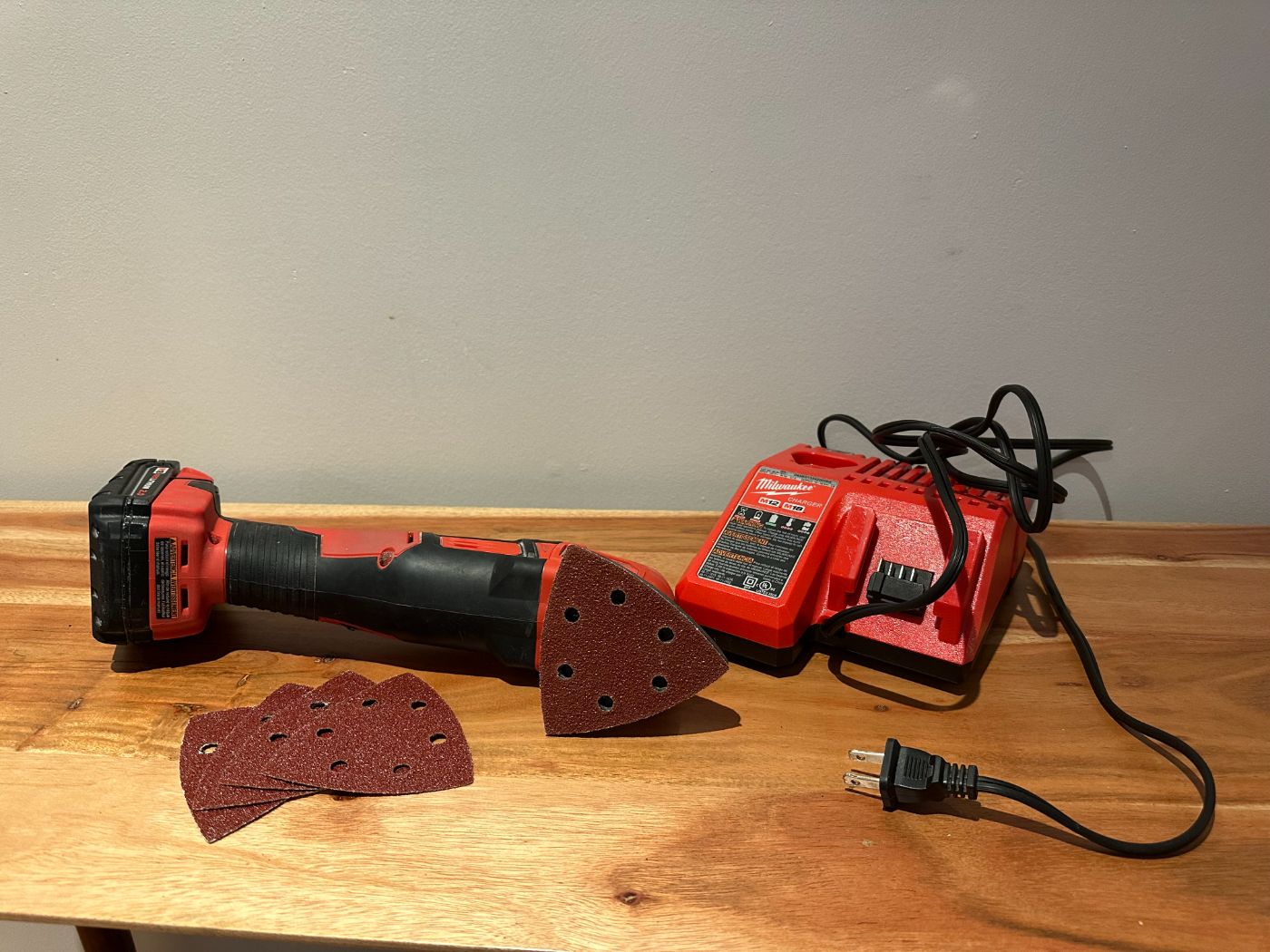 Ponceuse triangulaire sans fil Milwaukee avec chargeur et patins abrasifs grain 60, posée sur une table en bois