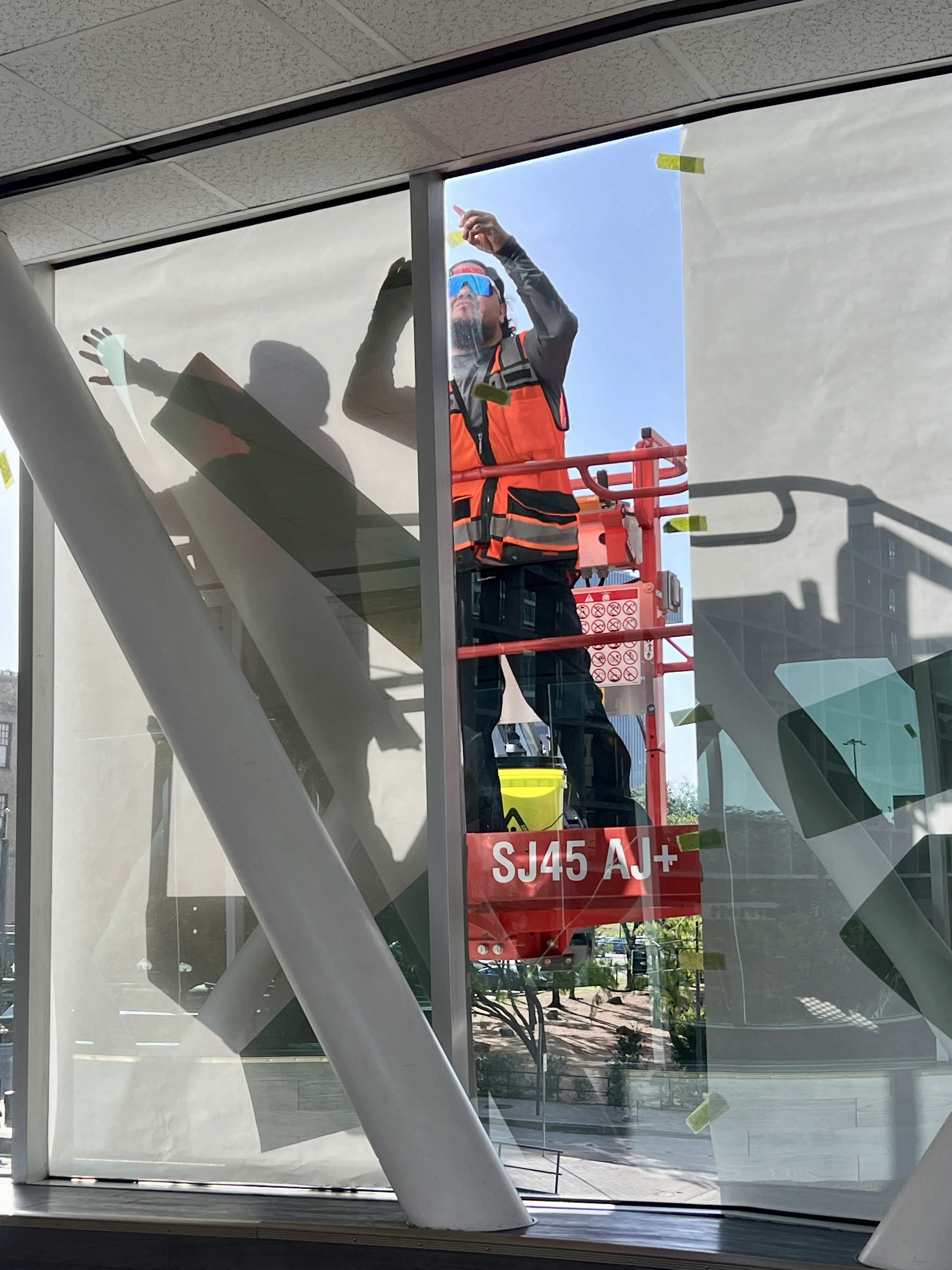 A worker in a bright orange vest uses a lift to reach high windows on a building under construction.