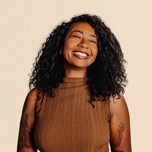 Smiling woman with curly hair wearing a sleeveless brown top, standing against a neutral background.