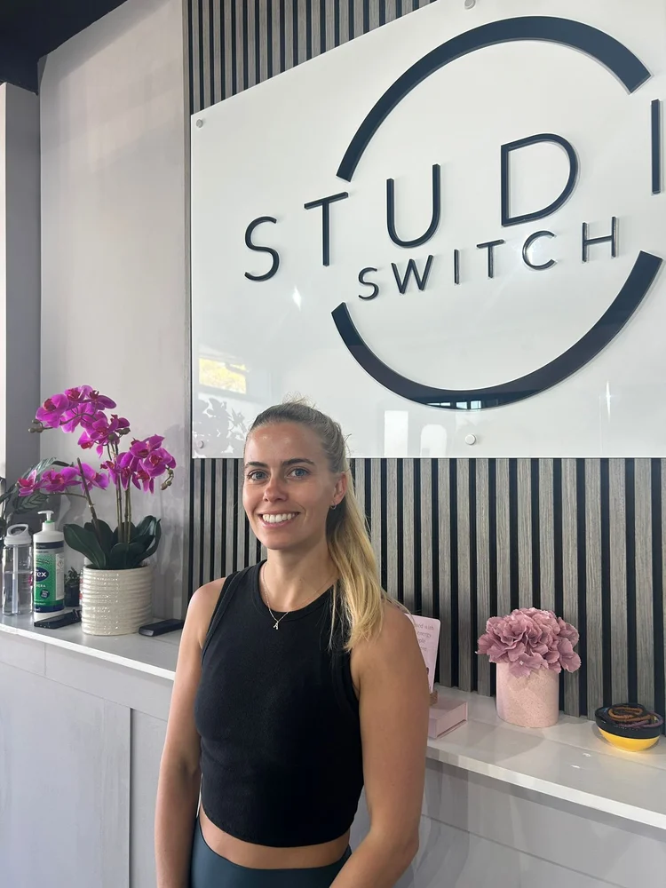 Amber, a Studio Switch Pilates instructor, seated at reception with orchids and the Studio Switch sign behind her.