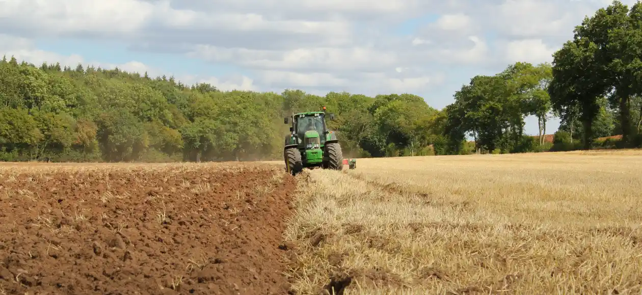 A green tractor plows a vast field under a partly cloudy sky. The tilled soil contrasts with the golden stubble, bordered by lush green trees.