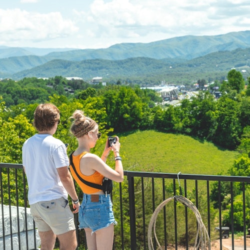 બાલ્કની પર બે લોકો હરિયાળાં લૅન્ડસ્કેપને જોઈ રહ્યા છે જેમાં વૃક્ષો અને દૂરના પર્વતો છે. એક વ્યક્તિ પાસે કેમેરા છે.