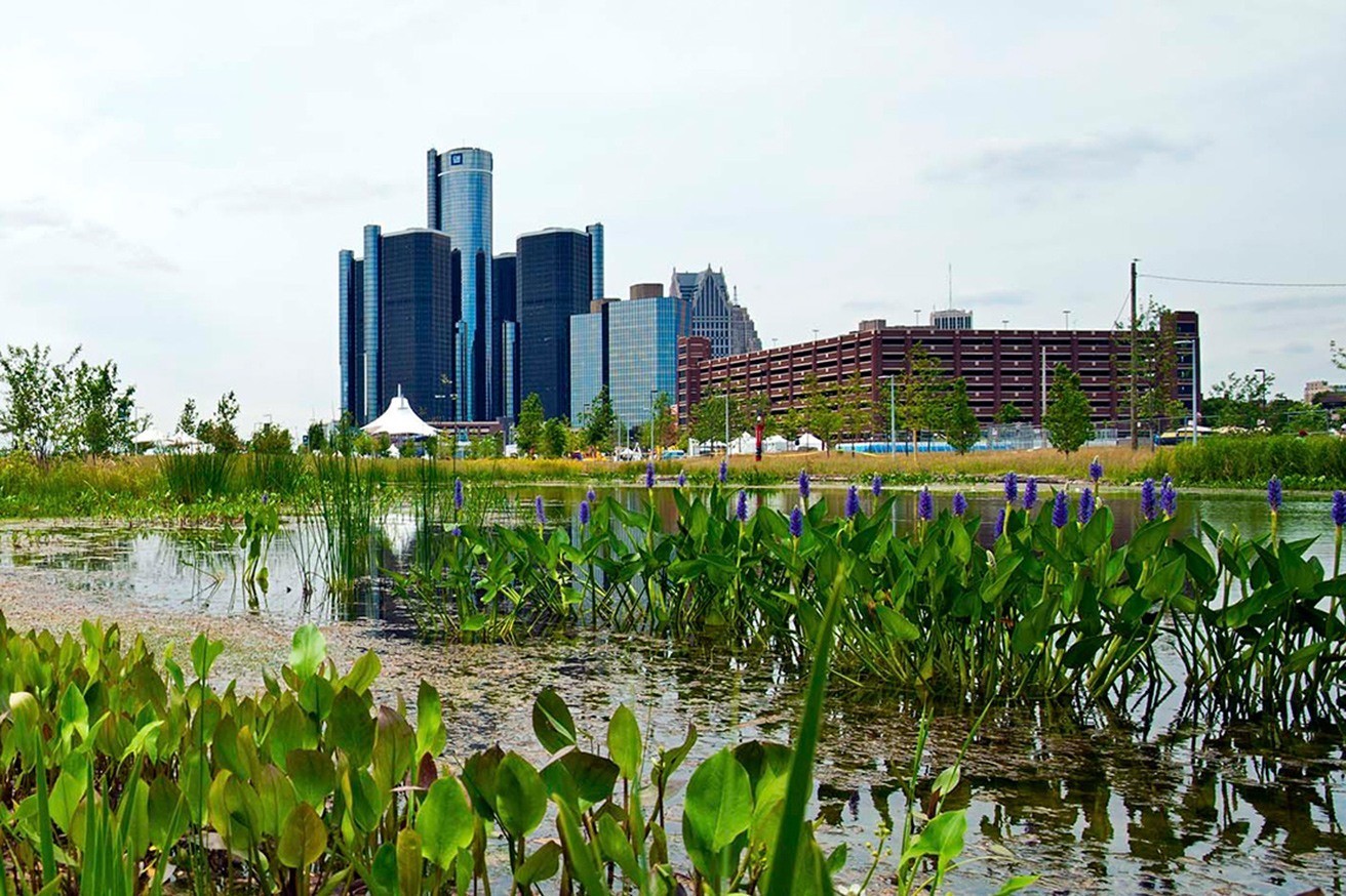 City skyline with modern buildings behind a reflective body of water and lush greenery in the foreground.