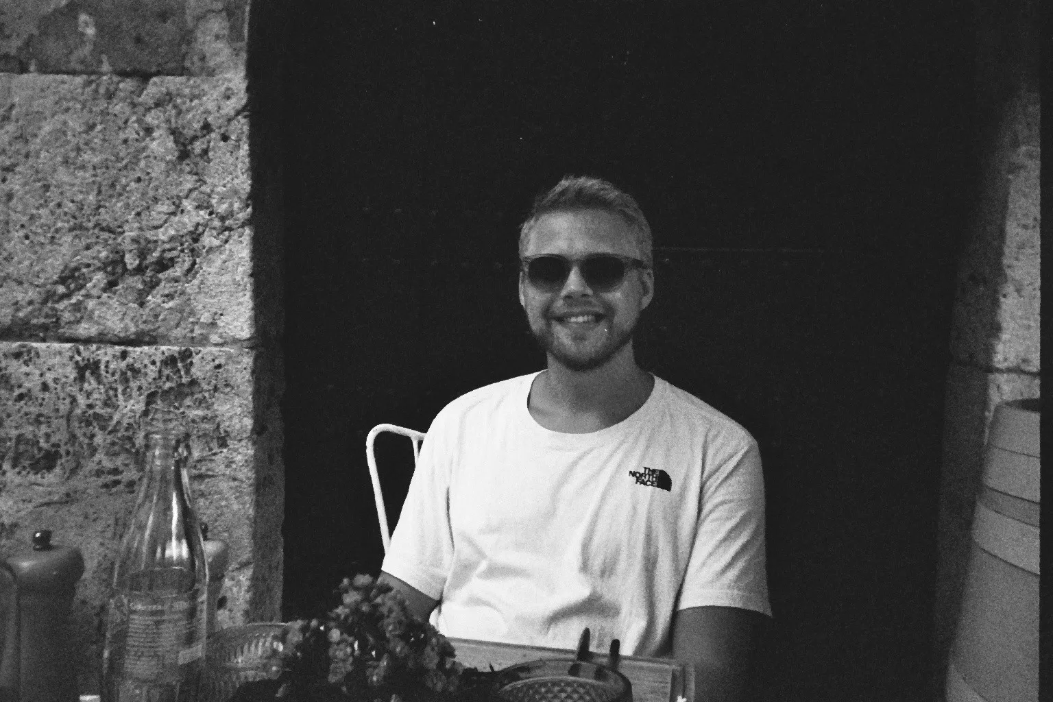 Man in sunglasses smiles while sitting at a table outdoors. He wears a t-shirt and looks at the camera.