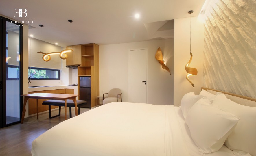 Wide-angle view of the spacious Echo Beach Suite studio in Canggu, Bali, showing the large bed with a textured accent wall, seating area, dining table, and kitchenette.