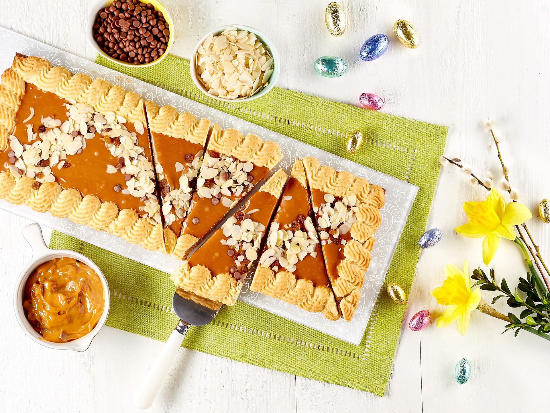 An overhead shot of a freshly baked caramel tart mazurek garnished with sliced almonds, displayed on a green placemat with colorful Easter eggs and a bouquet of yellow daffodils nearby, perfect for a food photo session.
