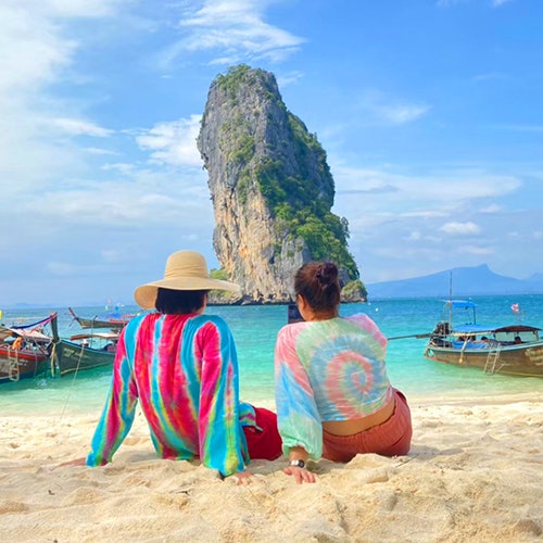 Two people in colorful shirts sit on a sandy beach facing the ocean, with boats and a tall rock formation in the background.