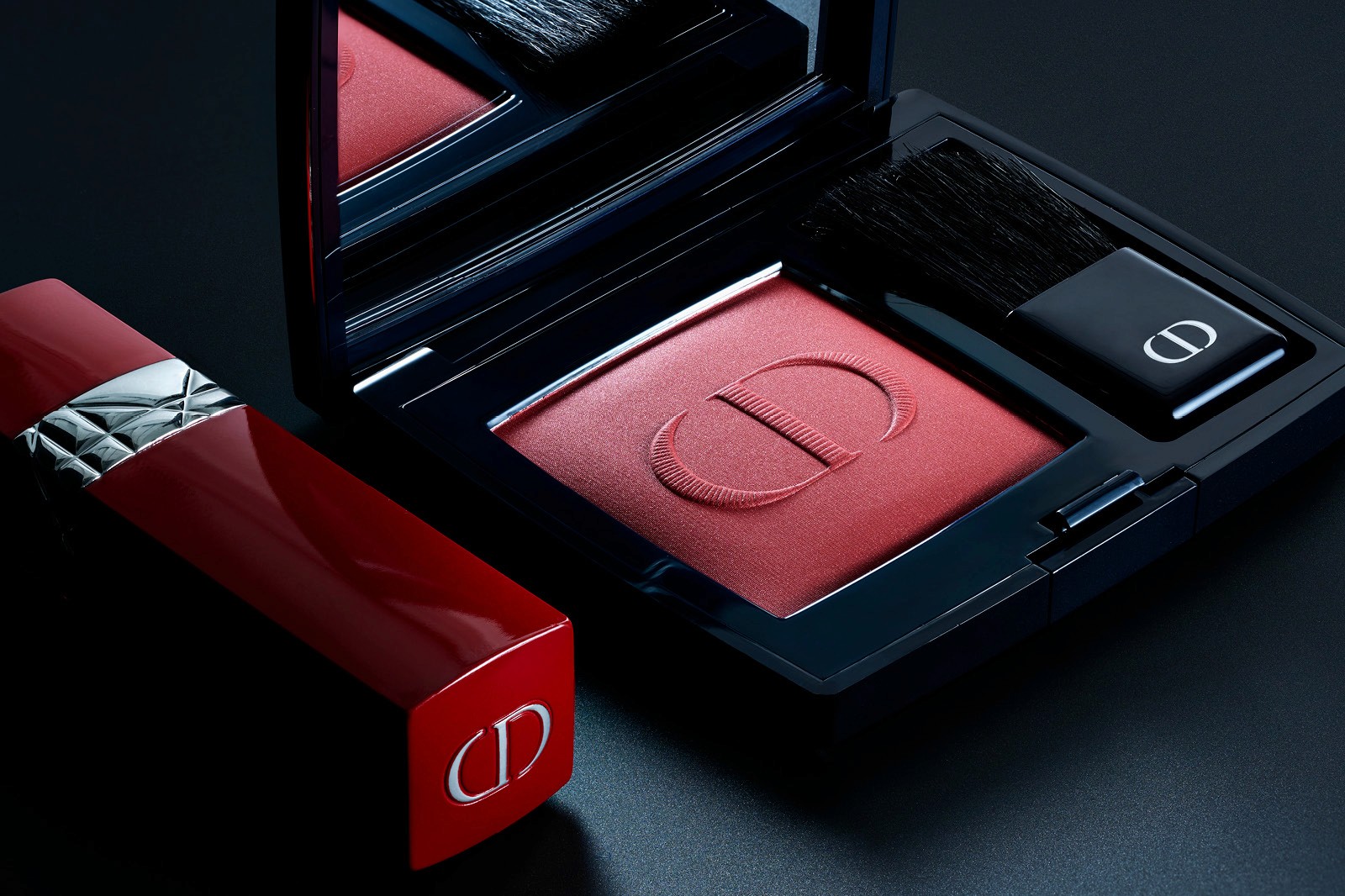 A black Christian Dior makeup compact with a red blush and a small applicator, next to a red lipstick tube on a dark background.