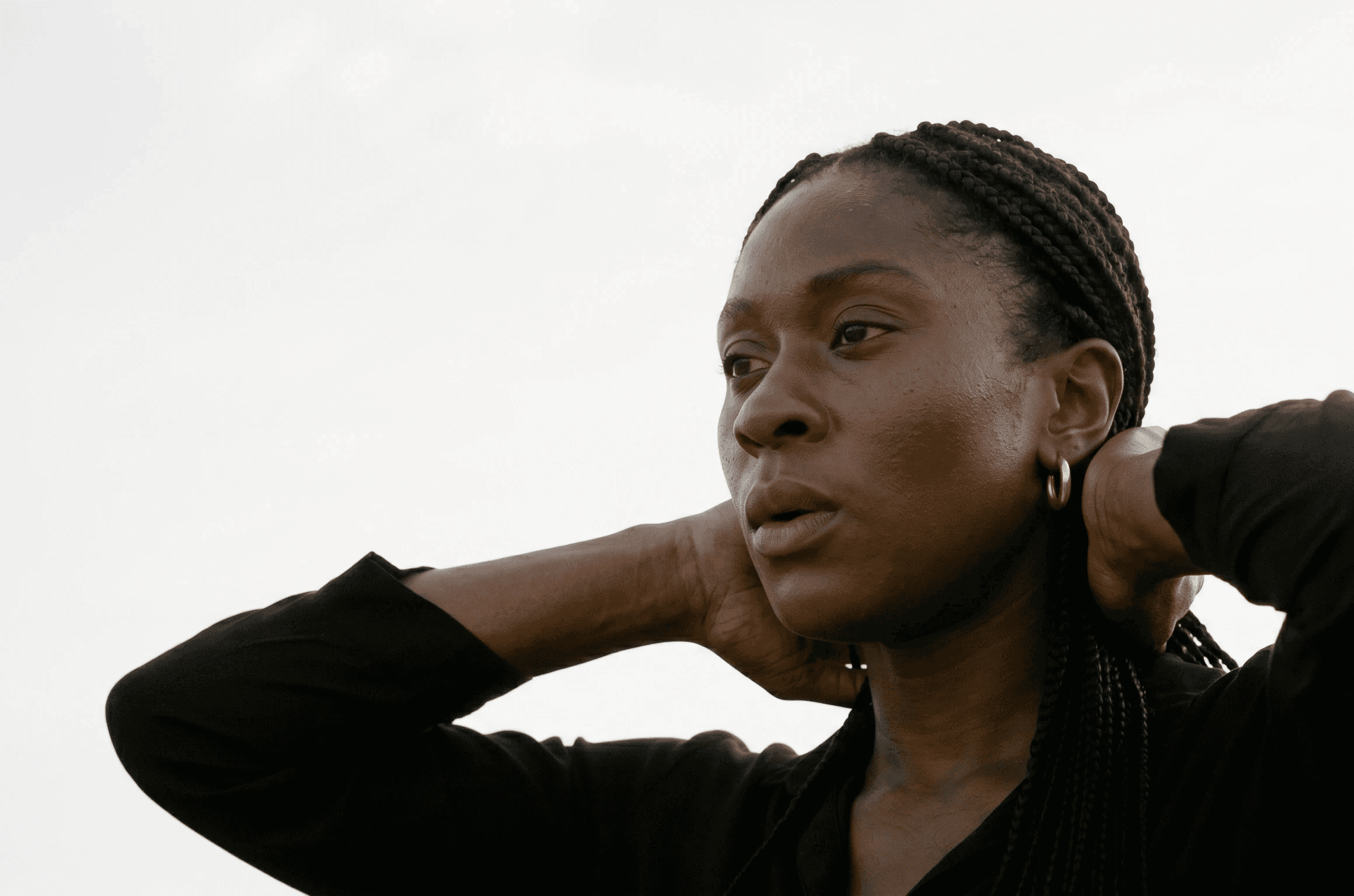 A person wearing a black top with braided hair is gazing thoughtfully off-camera against a neutral, pale background.