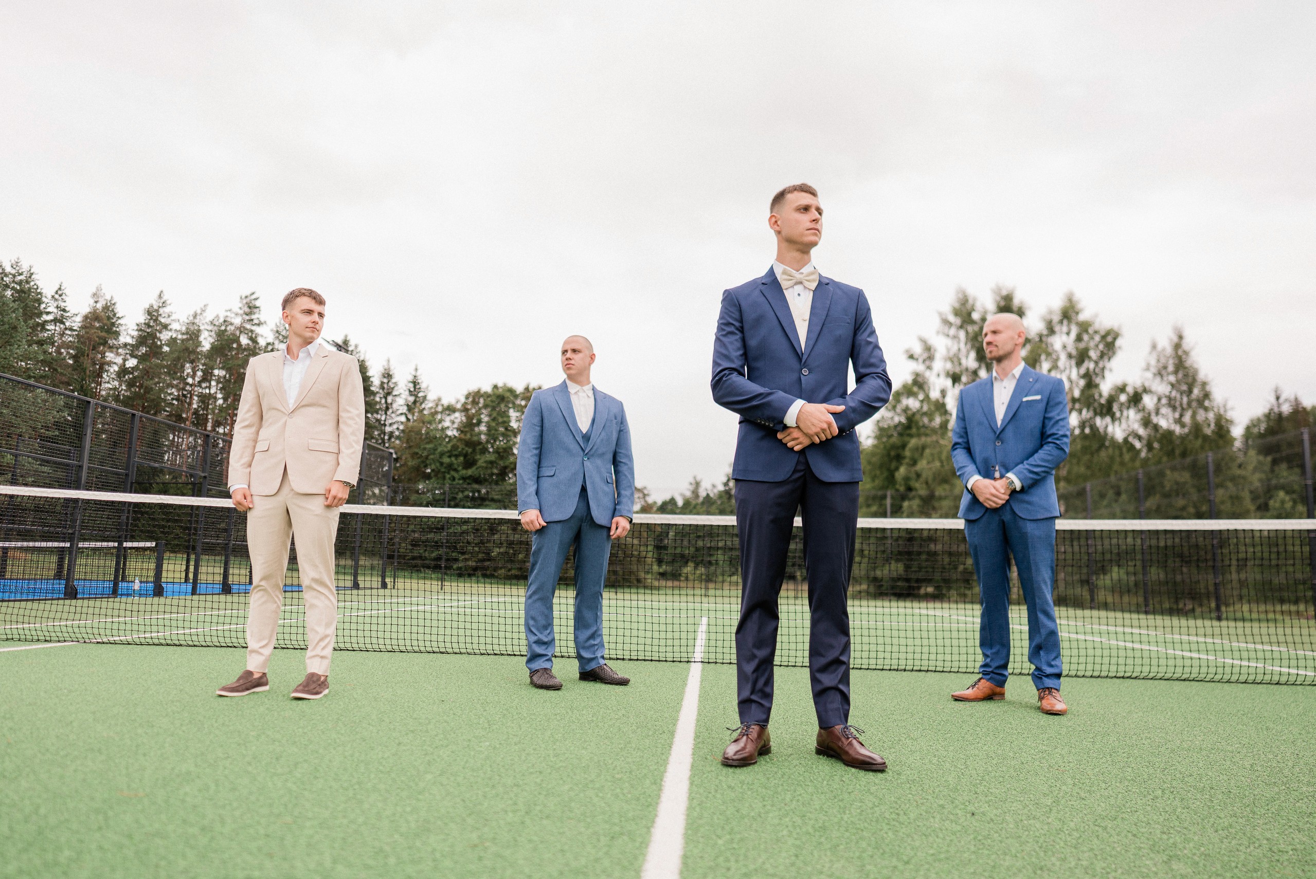 Four men in formal suits stand spaced across a tennis court, with the central groom positioned on the court line.