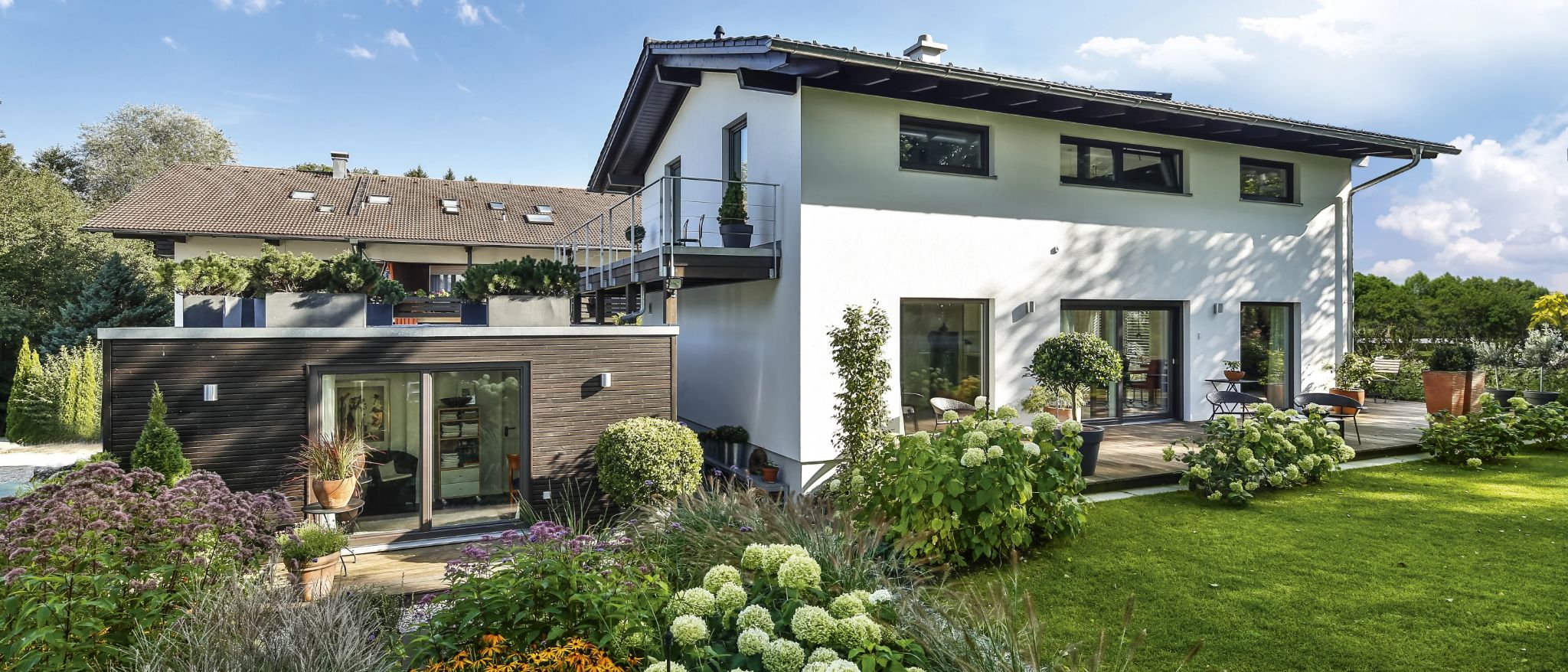 Moderne weiße Villa mit Holzakzenten, Balkon und gepflegtem Garten mit Gräsern, Blumen und Bäumen an einem sonnigen Tag.