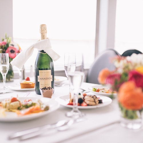 Mesa de comedor con platos de comida, una botella de champán Chandon, copas de vino y un ramo de flores coloridas.