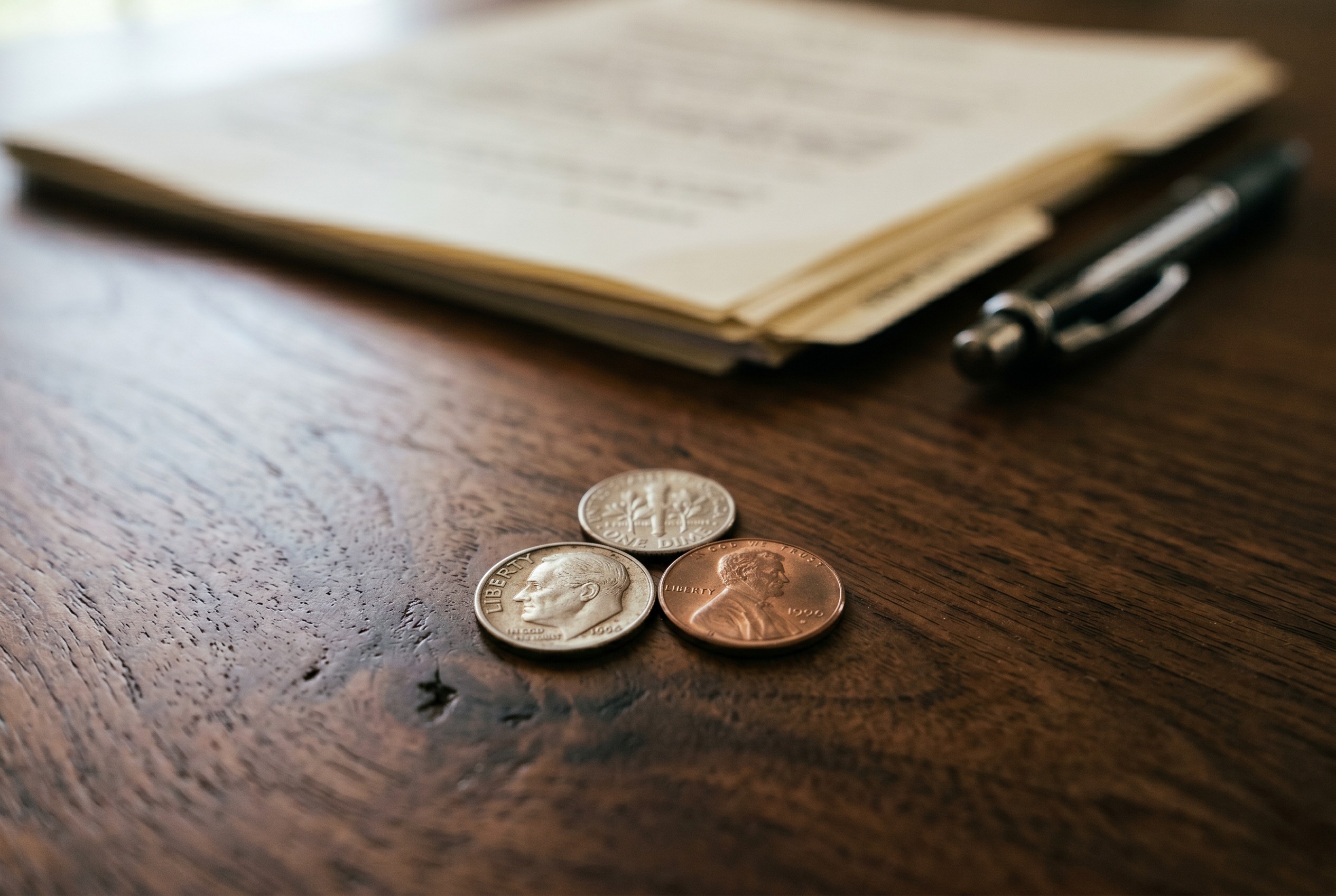 Two dimes and one penny totaling 21 cents on dark walnut desk with blurred legal documents and pen in background