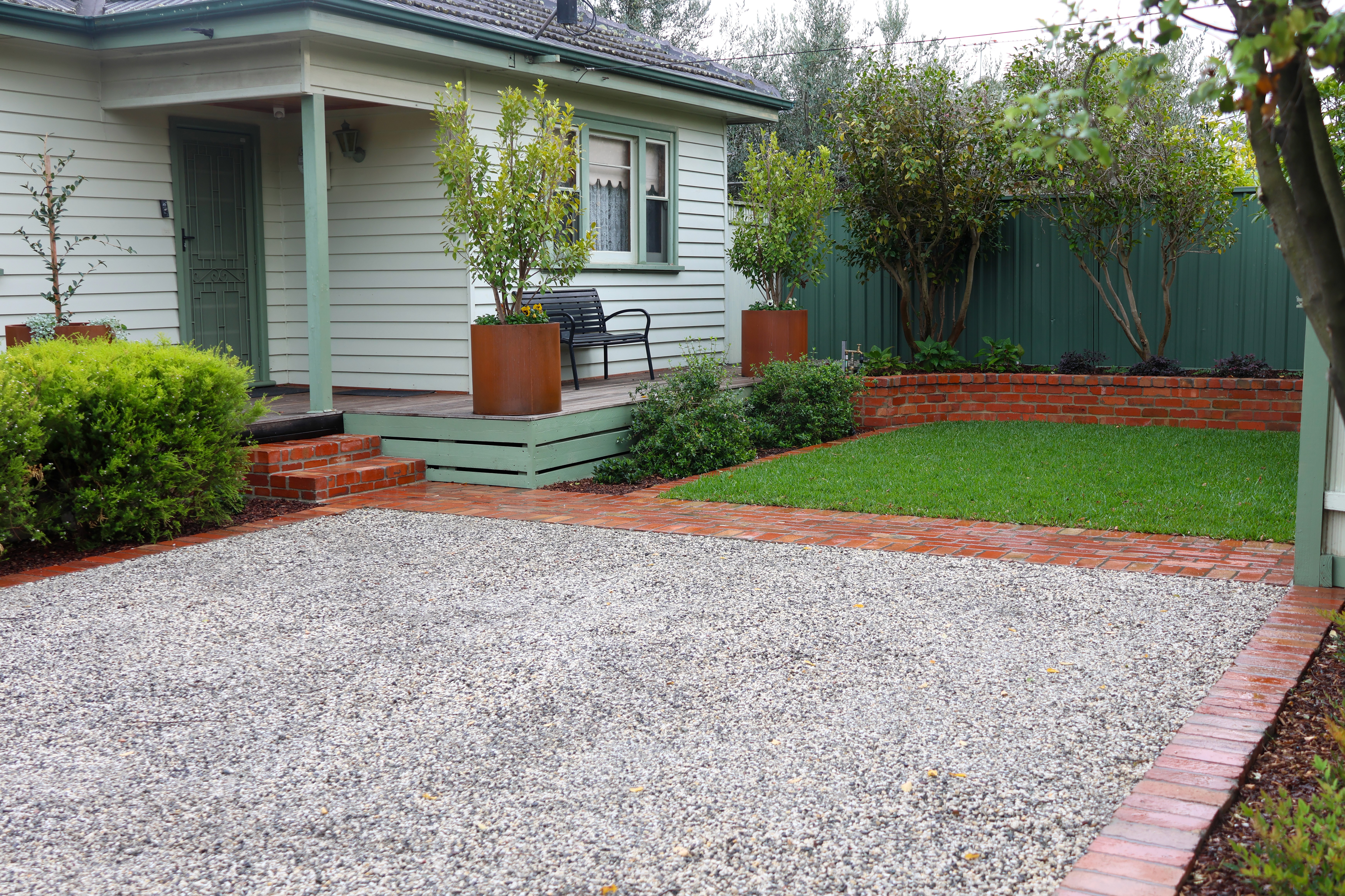 Full view of the renovated front yard of a 1950s weatherboard home featuring an exposed aggregate gravel driveway extension, a red brick pathway, lush green turf, and tall corten steel planters on the porch.