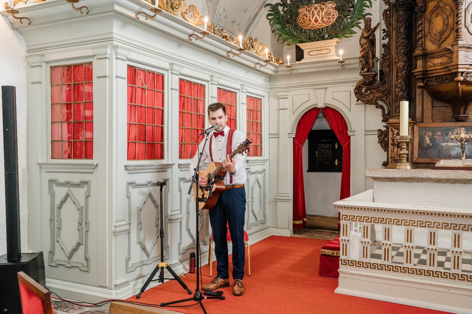 Ein Musiker spielt Akustikgitarre und singt in einer alten Kappele mit weißen Holzwänden und rotem Teppich.