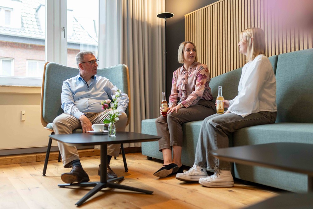 Zwei Geschäftsführer und eine Mitarbeiterin sitzen in Sesseln in einer Lounge-Ecke und sprechen miteinander in entspannter Atmosphäre.