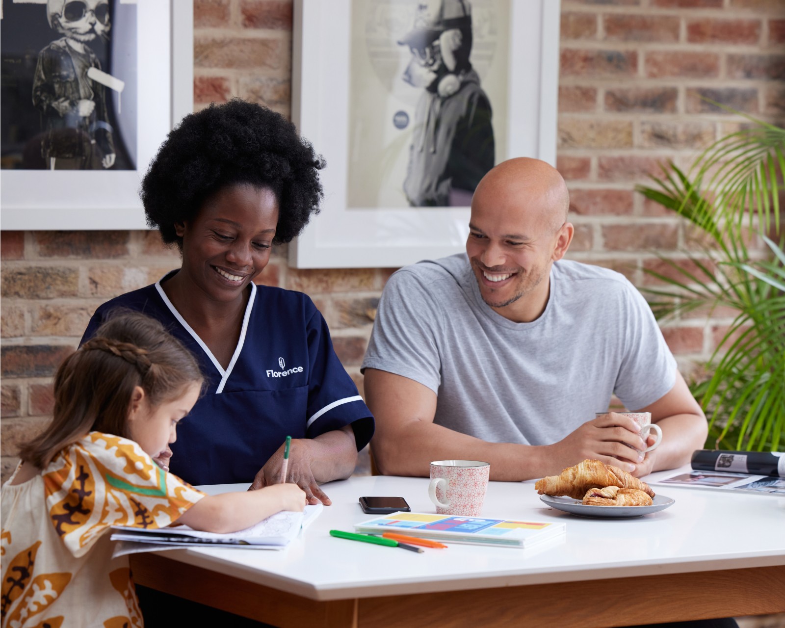 Caregiver in navy scrubs smiling while sitting at a table with a man and a child who is drawing, with coffee and pastries on the table in a home setting.