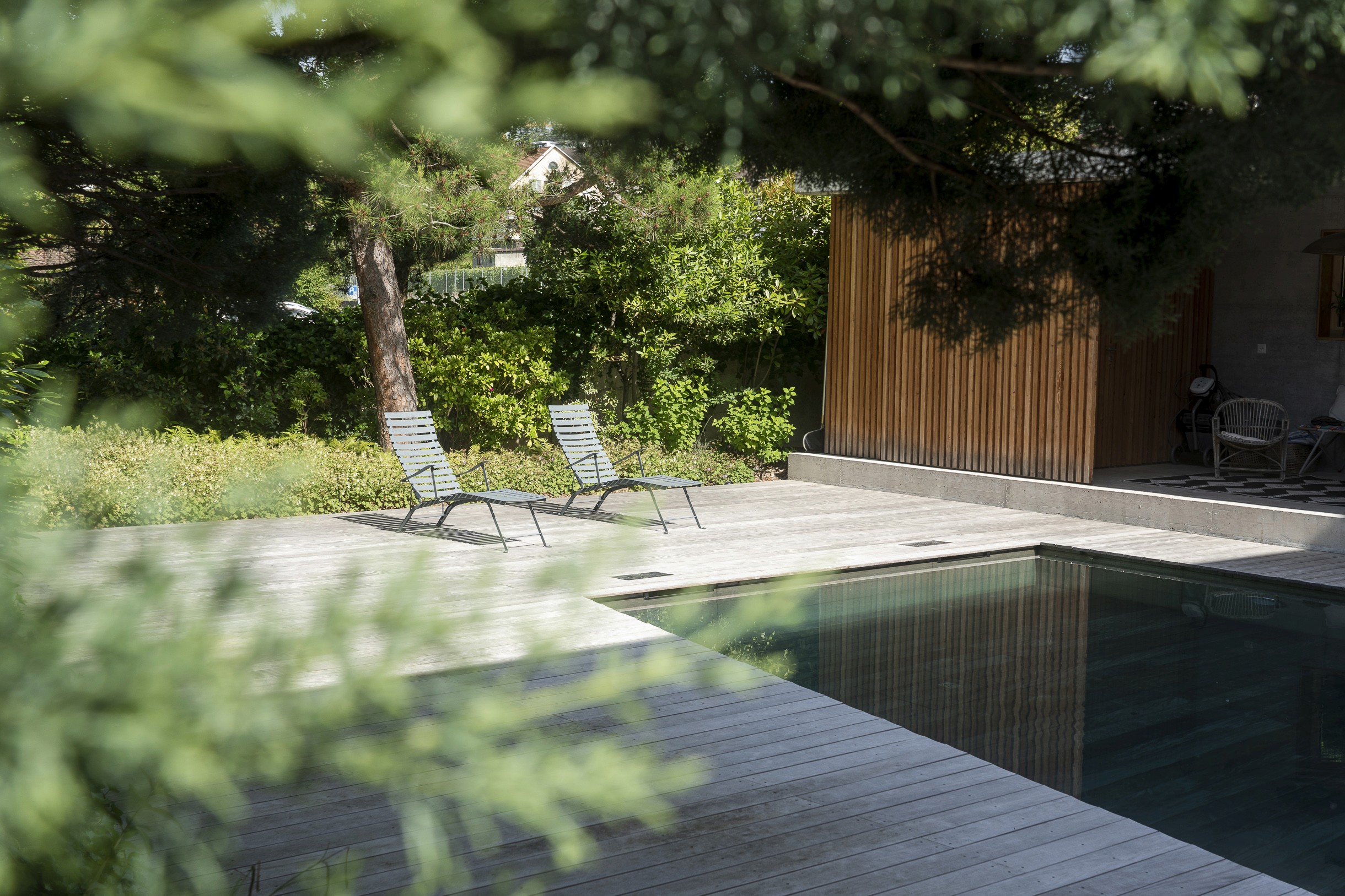 Photographie d'une réalisation du paysagiste Mirco Mancini chez un client privé montrant une terrace en bois autour d'une piscine avec deux chaises longues et ombragée par des arbres