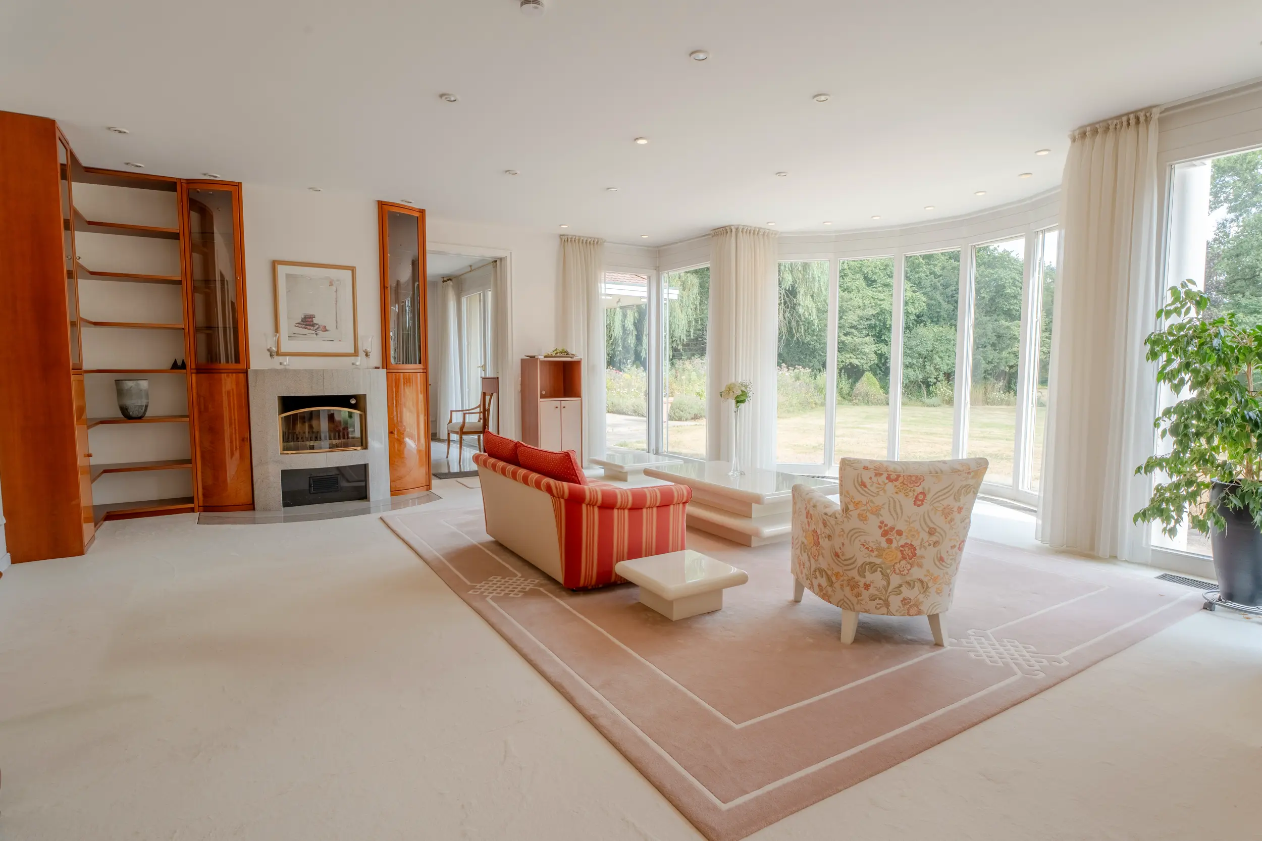 Elegantes Wohnzimmer der Villa Gronau mit offenem Kamin, Einbauregalen und Panoramafenstern zum Garten – Westfalen Wohnen"