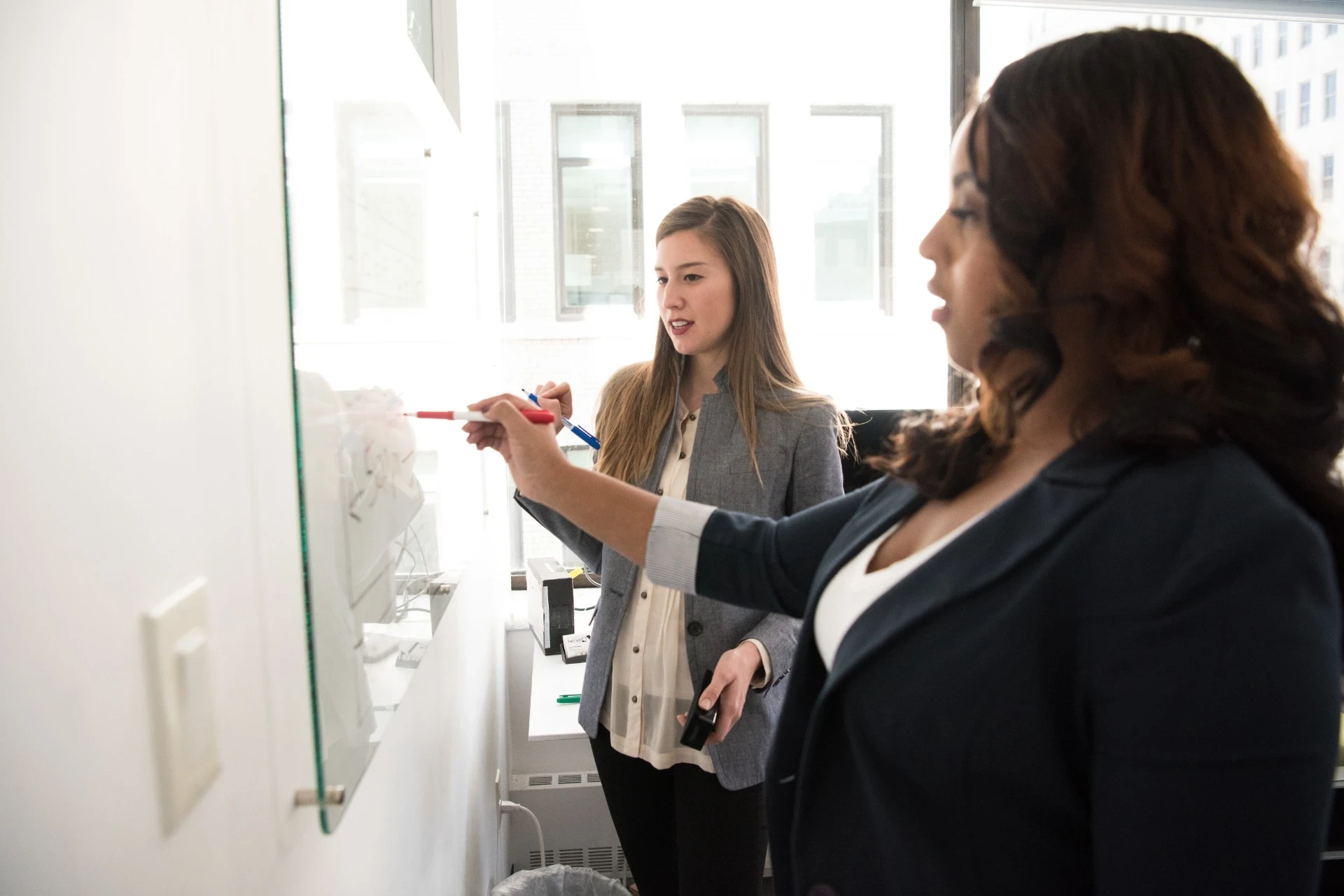 Imagem de duas mulheres em um ambiente corporativo colaborando em uma apresentação. Elas estão em pé, escrevendo em uma lousa ou vidro transparente, utilizando marcadores de cores diferentes. Ambas estão vestidas de forma profissional e focadas na tarefa. A mulher em segundo plano observa enquanto a outra escreve, demonstrando um momento de trabalho em equipe e troca de ideias. A cena ocorre em uma sala iluminada, com janelas que permitem a entrada de luz natural.