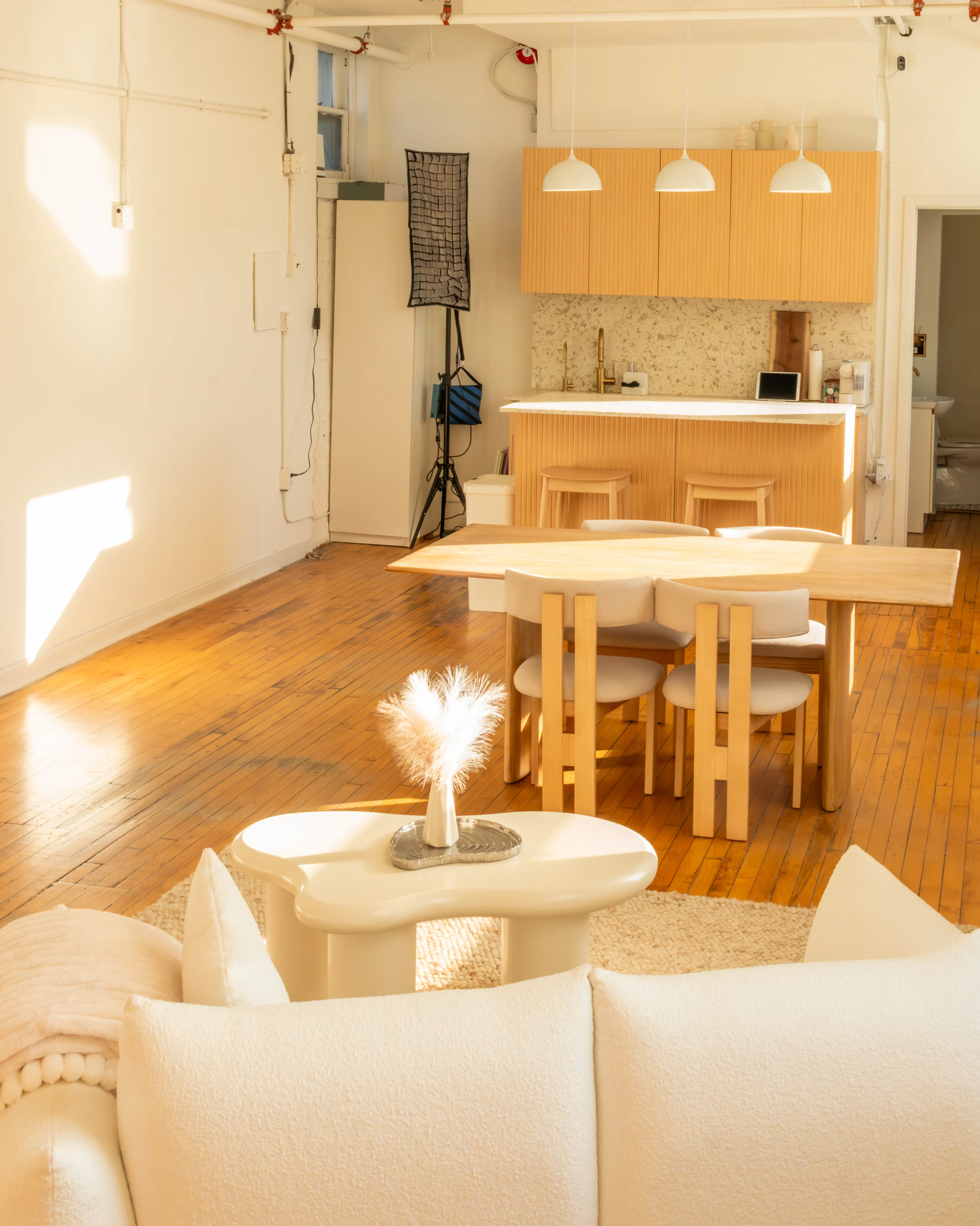 Wide view of Node Studio showcasing its open-concept layout with a cozy white couch, a minimalist dining area, hardwood floors, and a semi-equipped kitchen, all bathed in abundant natural light