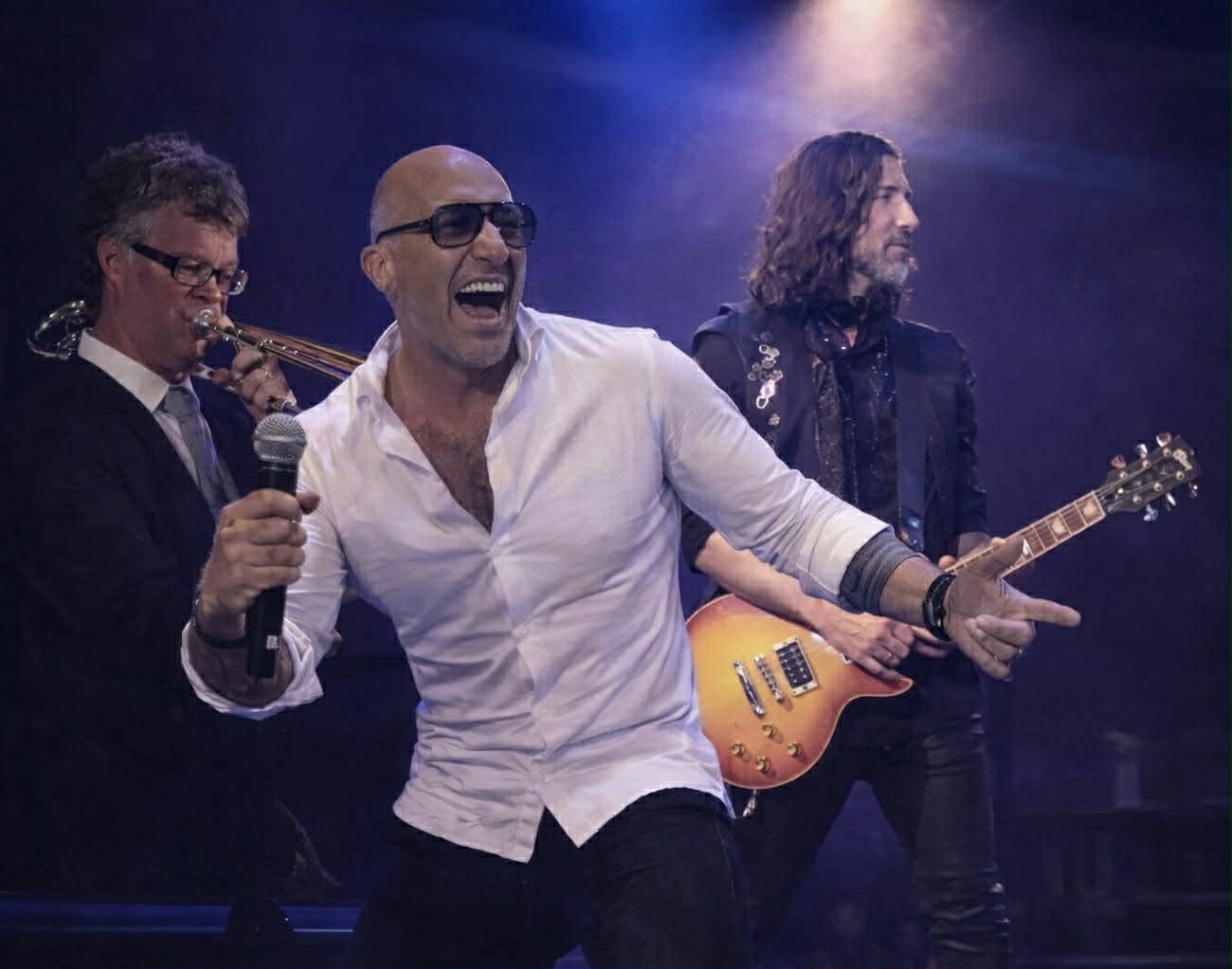 A young person with curly hair smiles at the camera while sitting in front of a laptop against a dark background.
