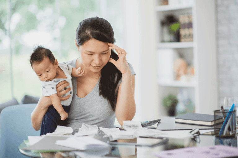 Mother holding a baby while looking stressed over bills and paperwork.