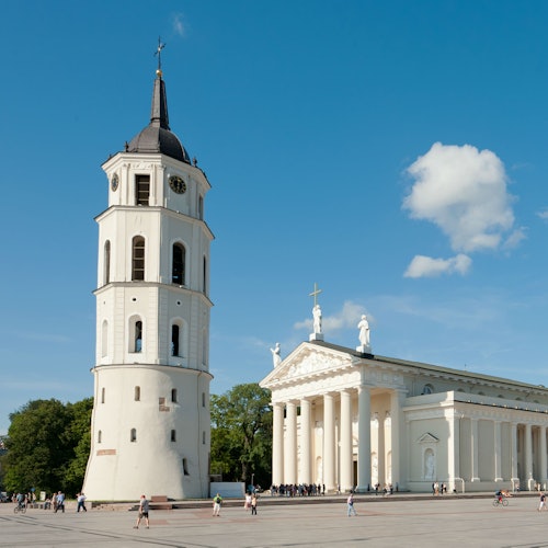 A historic tower and a white neoclassical building with columns under a clear blue sky, people walking in the plaza below.