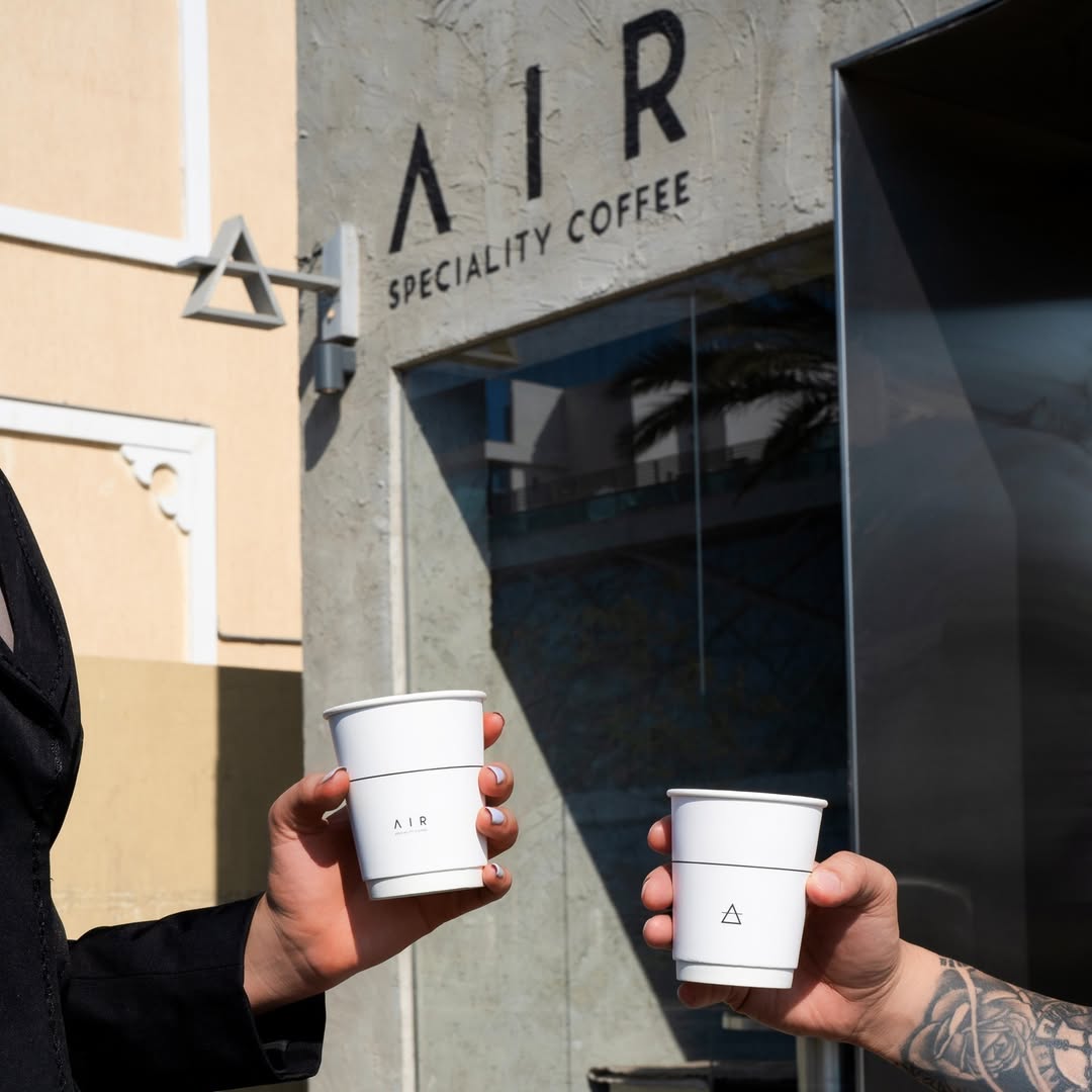 Two people holding coffee cups in front of the Air Specialty Coffee shop board.