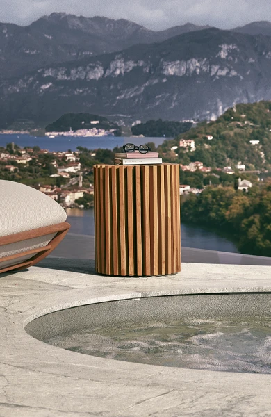 Round Layer side table in solid Iroko wood, composed of interlocked vertical panels in a radial pattern, styled in a luxury poolside setting with lounge chairs and tropical greenery – modern outdoor furniture with sculptural design.