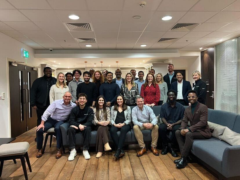 A photo of Sam and his colleagues. Some sat on a long L shaped couch and others standing directly behind. Everyone is smiling :).