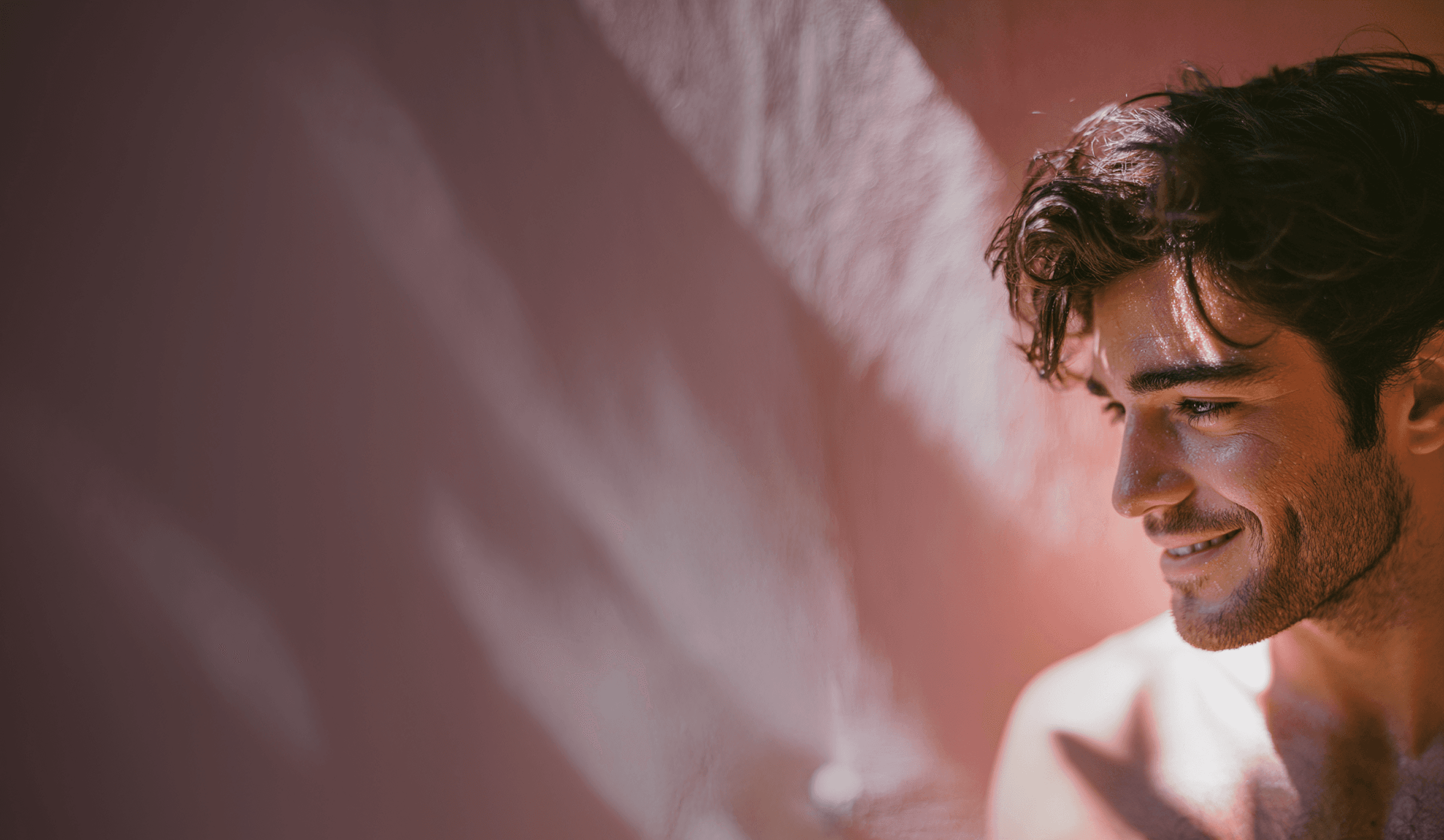 Smiling man with curly hair in soft lighting against a pink wall. The image conveys a warm and relaxed tone with natural shadows.