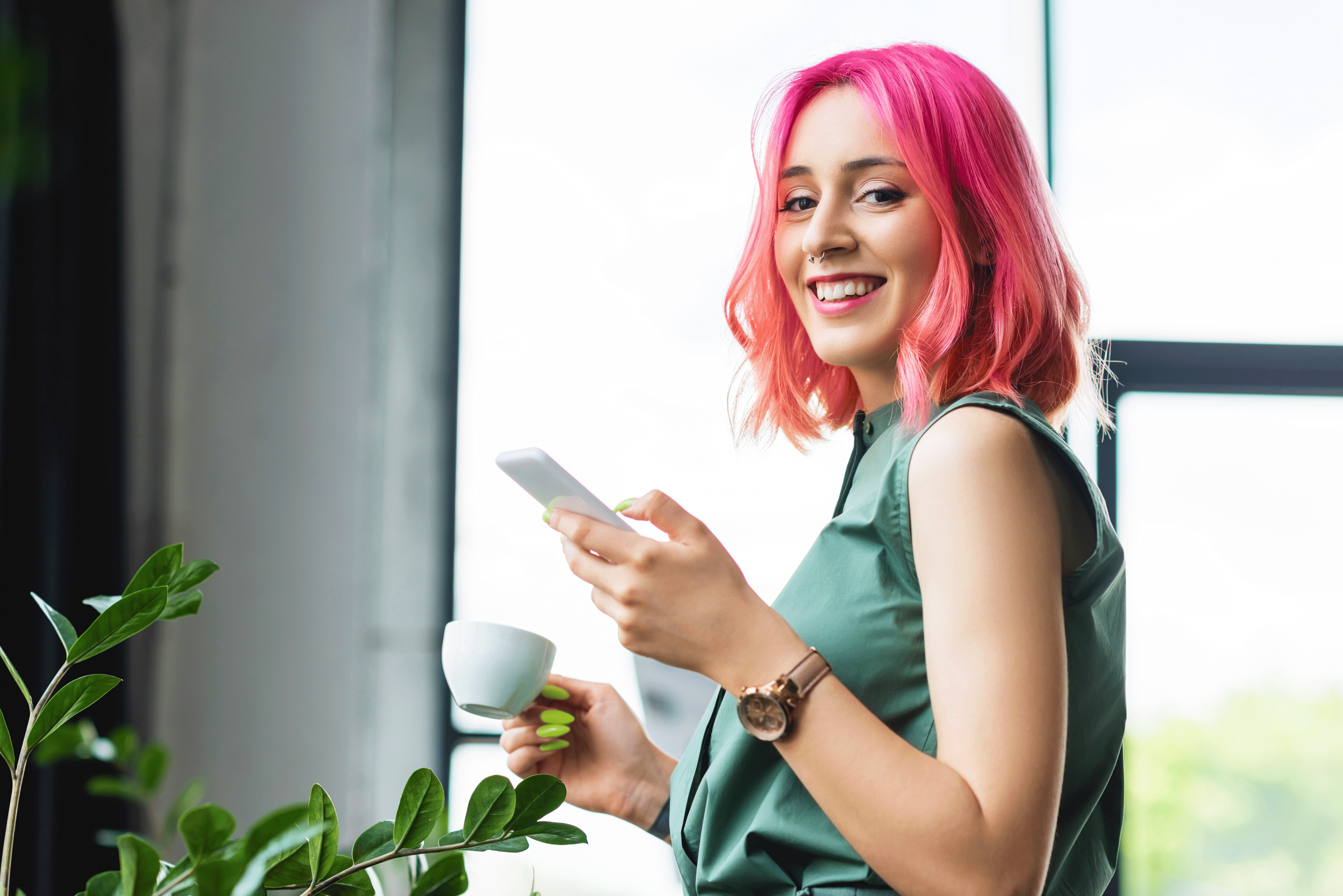 A smiling woman with bright pink hair and a septum piercing looks toward the camera while holding a smartphone and a small white coffee cup. She is wearing a green sleeveless top and a wristwatch, standing in a bright indoor space with green plants in the foreground.